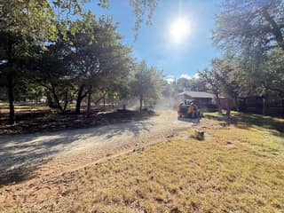 The skid steer grading the old gravel driveway in a cloud of dust, with the carport in the background