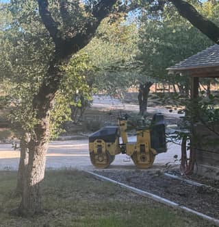 A yellow vibratory roller compacting the road base under the trees near the house